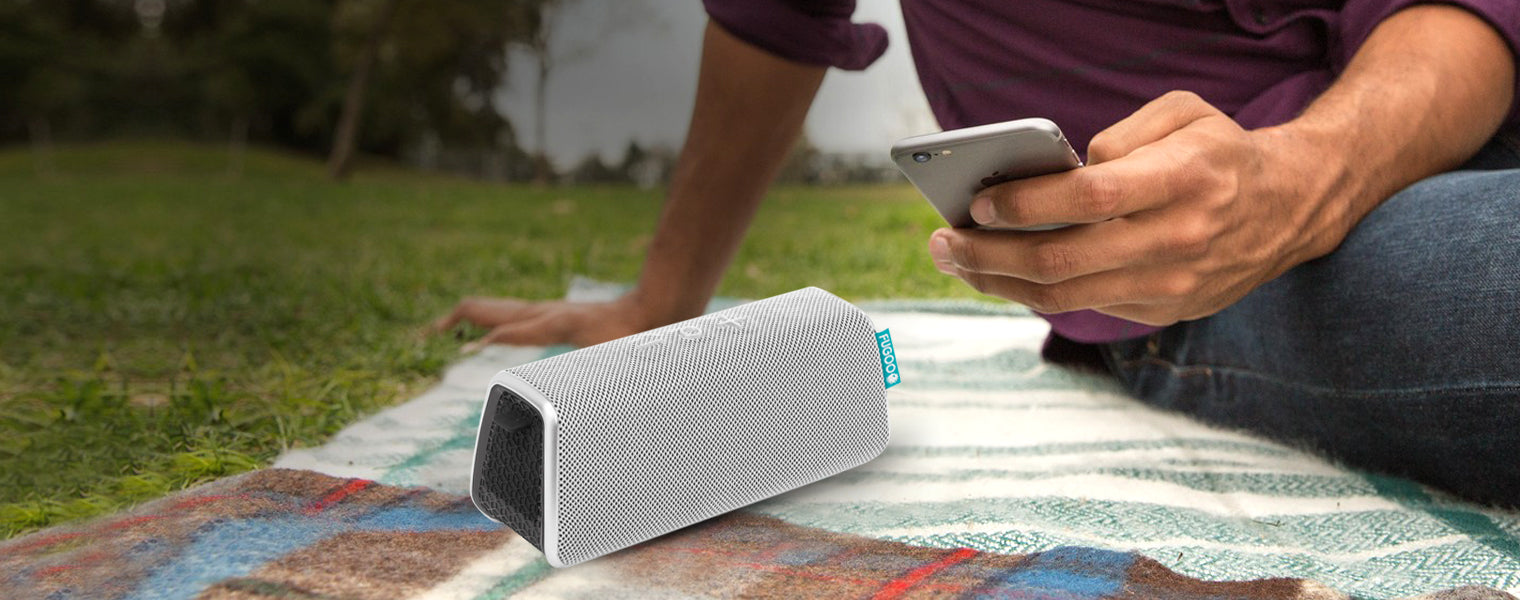 Person in purple shirt sitting on a patterned picnic blanket in grassy park, using smartphone beside a white portable Bluetooth speaker with teal accent, demonstrating outdoor connectivity use