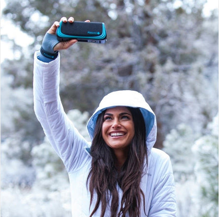Woman in white winter clothing and sun hat taking a selfie with a teal phone case against a snowy background, smiling broadly during outdoor winter adventure