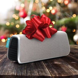 White portable Bluetooth speaker with red gift bow on wooden surface against blurred Christmas tree lights background, perfect for holiday gifting