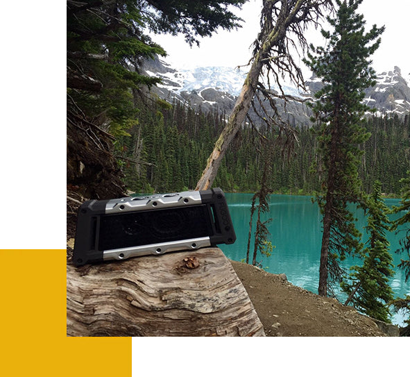 A black rugged speaker rests on a log beside a turquoise mountain lake and towering evergreens.